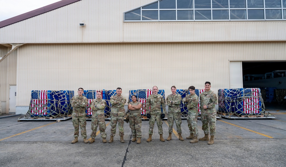 A group of nine military personnel in MultiCam/OCP camouflage uniforms pose in front of cargo pallets wrapped with American flag-patterned coverings and secured with cargo nets. They are standing on a tarmac or flight line apron in front of a large military hangar facility. The group appears to be posing for an official or commemorative photograph.
