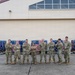 A group of nine military personnel in MultiCam/OCP camouflage uniforms pose in front of cargo pallets wrapped with American flag-patterned coverings and secured with cargo nets. They are standing on a tarmac or flight line apron in front of a large military hangar facility. The group appears to be posing for an official or commemorative photograph.