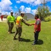 Military personnel in camouflage uniforms and high-visibility safety vests are engaged in what appears to be disaster relief or recovery operations. A soldier wearing a white hard hat and reflective vest with a Corps of Engineers-style insignia is shaking hands with a civilian in a red shirt. The surrounding environment shows storm-damaged trees and structures, suggesting post-hurricane or typhoon recovery work.