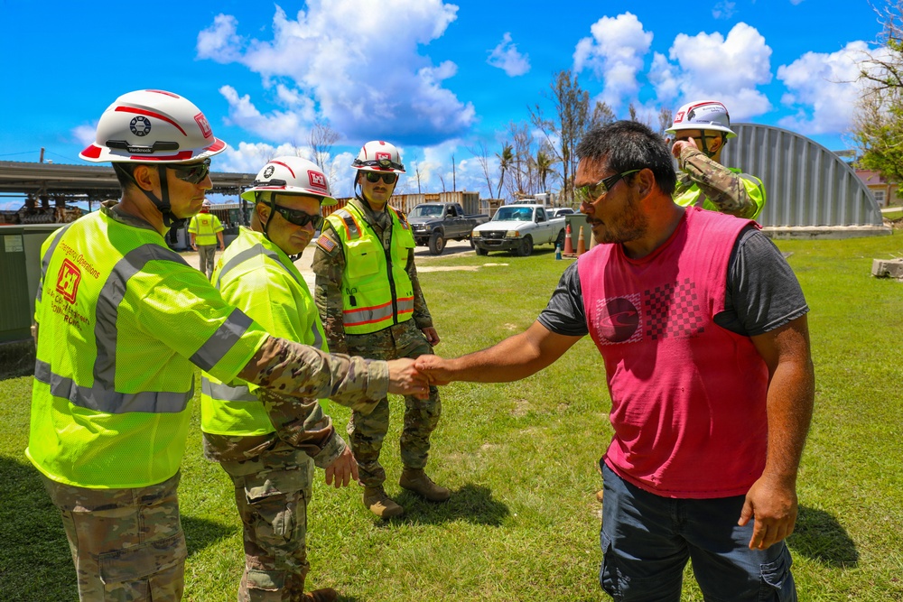 Military personnel and emergency operations workers in high-visibility vests and white hard hats are greeting a civilian man wearing a red vest in an outdoor area showing signs of storm or disaster damage. A handshake is taking place between a soldier in camouflage uniform and the civilian, suggesting a coordination or relief effort meeting. The background shows stripped or damaged trees, vehicles, and a metal structure consistent with post-disaster recovery operations.