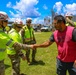 Military personnel and emergency operations workers in high-visibility vests and white hard hats are greeting a civilian man wearing a red vest in an outdoor area showing signs of storm or disaster damage. A handshake is taking place between a soldier in camouflage uniform and the civilian, suggesting a coordination or relief effort meeting. The background shows stripped or damaged trees, vehicles, and a metal structure consistent with post-disaster recovery operations.