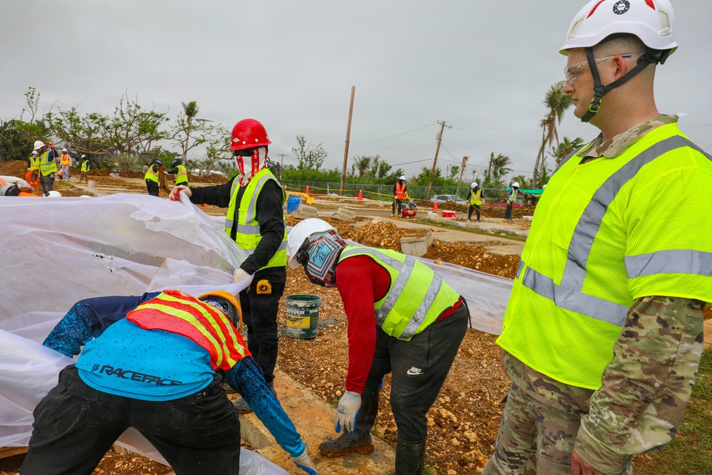 A disaster recovery or construction operation is underway at a site showing significant storm damage, with stripped and downed trees visible in the background. Workers in high-visibility safety vests and hard hats are actively working at ground level, while a military personnel member in MultiCam/OCP uniform and white hard hat observes the operation. The scene suggests post-hurricane or major storm recovery efforts, likely involving infrastructure reconstruction.