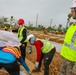 A disaster recovery or construction operation is underway at a site showing significant storm damage, with stripped and downed trees visible in the background. Workers in high-visibility safety vests and hard hats are actively working at ground level, while a military personnel member in MultiCam/OCP uniform and white hard hat observes the operation. The scene suggests post-hurricane or major storm recovery efforts, likely involving infrastructure reconstruction.