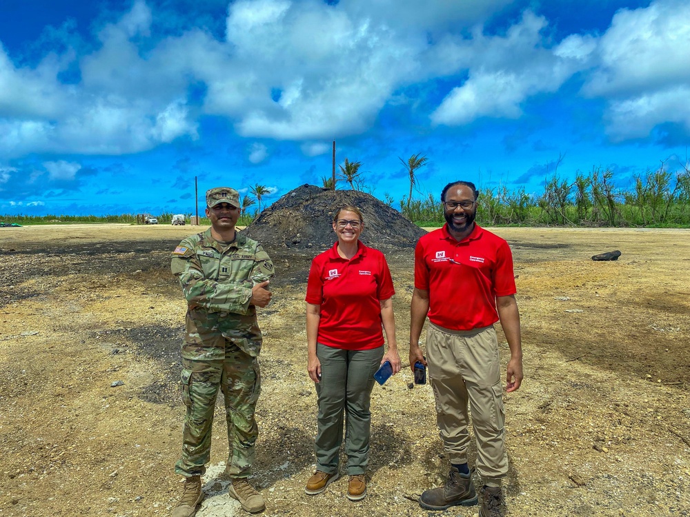 Three individuals stand together outdoors at what appears to be a disaster recovery or debris management site. One person is wearing U.S. Army military camouflage uniform, while the other two wear matching red polo shirts with logos consistent with emergency management or relief organization branding. The background shows storm-damaged vegetation, a large mound of debris, and cleared ground, suggesting post-disaster cleanup operations.