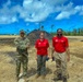Three individuals stand together outdoors at what appears to be a disaster recovery or debris management site. One person is wearing U.S. Army military camouflage uniform, while the other two wear matching red polo shirts with logos consistent with emergency management or relief organization branding. The background shows storm-damaged vegetation, a large mound of debris, and cleared ground, suggesting post-disaster cleanup operations.