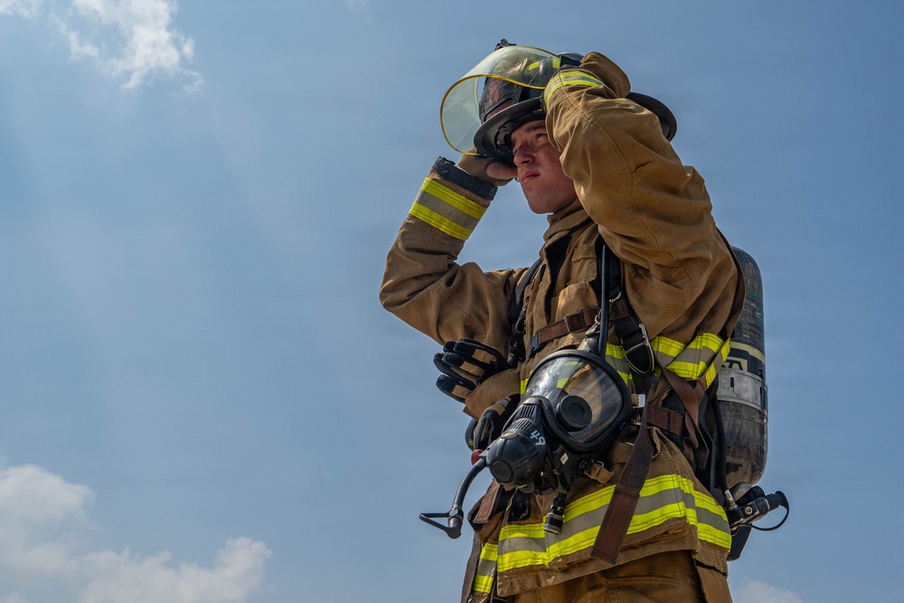A firefighter in full turnout gear is photographed from a low angle against a partly cloudy blue sky. The individual is adjusting their helmet visor with one gloved hand while carrying full SCBA (Self-Contained Breathing Apparatus) equipment on their back. The image captures a moment of preparation or post-incident activity.