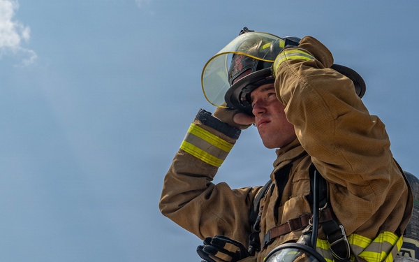 A firefighter in full turnout gear is photographed from a low angle against a partly cloudy blue sky. The individual is adjusting their helmet visor with one gloved hand while carrying full SCBA (Self-Contained Breathing Apparatus) equipment on their back. The image captures a moment of preparation or post-incident activity.