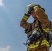 A firefighter in full turnout gear is photographed from a low angle against a partly cloudy blue sky. The individual is adjusting their helmet visor with one gloved hand while carrying full SCBA (Self-Contained Breathing Apparatus) equipment on their back. The image captures a moment of preparation or post-incident activity.