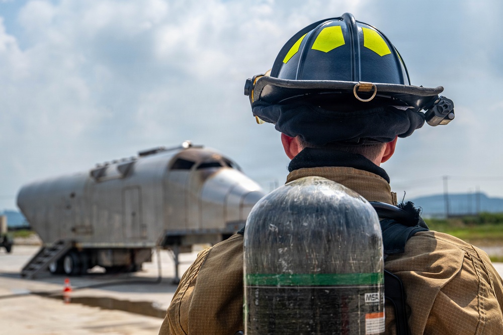 A firefighter in full protective gear, including a black helmet with yellow-green reflective markings and an MSA SCBA (Self-Contained Breathing Apparatus) tank, stands on a tarmac facing a mock aircraft fuselage mounted on a trailer. The scene appears to be an aircraft rescue and firefighting (ARFF) training exercise at an airfield. The firefighter is viewed from behind, observing the training prop in the distance.