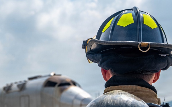 A firefighter in full protective gear, including a black helmet with yellow-green reflective markings and an MSA SCBA (Self-Contained Breathing Apparatus) tank, stands on a tarmac facing a mock aircraft fuselage mounted on a trailer. The scene appears to be an aircraft rescue and firefighting (ARFF) training exercise at an airfield. The firefighter is viewed from behind, observing the training prop in the distance.