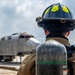 A firefighter in full protective gear, including a black helmet with yellow-green reflective markings and an MSA SCBA (Self-Contained Breathing Apparatus) tank, stands on a tarmac facing a mock aircraft fuselage mounted on a trailer. The scene appears to be an aircraft rescue and firefighting (ARFF) training exercise at an airfield. The firefighter is viewed from behind, observing the training prop in the distance.