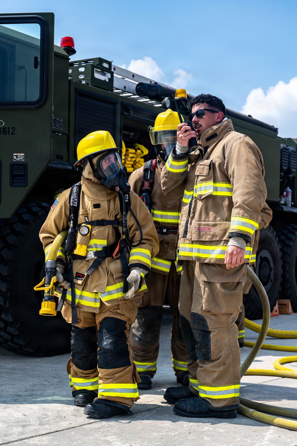 Three firefighters in full turnout gear stand in front of a military-style olive green fire apparatus (vehicle number 3612). One firefighter on the left is fully equipped with SCBA (Self-Contained Breathing Apparatus) and a full-face mask, holding a hose nozzle. The firefighter on the right is communicating via handheld radio while holding a fire hose, suggesting an active training exercise or operational scenario.