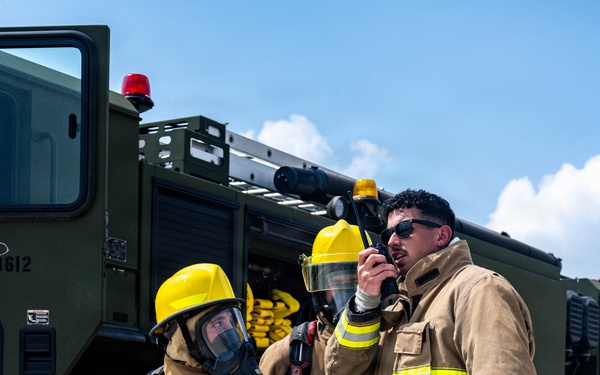 Three firefighters in full turnout gear stand in front of a military-style olive green fire apparatus (vehicle number 3612). One firefighter on the left is fully equipped with SCBA (Self-Contained Breathing Apparatus) and a full-face mask, holding a hose nozzle. The firefighter on the right is communicating via handheld radio while holding a fire hose, suggesting an active training exercise or operational scenario.