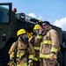 Three firefighters in full turnout gear stand in front of a military-style olive green fire apparatus (vehicle number 3612). One firefighter on the left is fully equipped with SCBA (Self-Contained Breathing Apparatus) and a full-face mask, holding a hose nozzle. The firefighter on the right is communicating via handheld radio while holding a fire hose, suggesting an active training exercise or operational scenario.