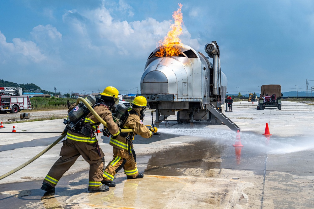 Two firefighters in full protective gear are conducting what appears to be an aircraft rescue and firefighting (ARFF) training exercise. They are directing foam/water suppression agent from a hose at a mock aircraft fuselage simulator that has active flames burning from its top. The exercise is taking place on a paved tarmac-like surface with support vehicles and equipment visible in the background.