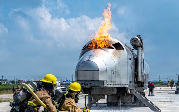 Two firefighters in full protective gear are conducting what appears to be an aircraft rescue and firefighting (ARFF) training exercise. They are directing foam/water suppression agent from a hose at a mock aircraft fuselage simulator that has active flames burning from its top. The exercise is taking place on a paved tarmac-like surface with support vehicles and equipment visible in the background.