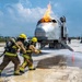 Two firefighters in full protective gear are conducting what appears to be an aircraft rescue and firefighting (ARFF) training exercise. They are directing foam/water suppression agent from a hose at a mock aircraft fuselage simulator that has active flames burning from its top. The exercise is taking place on a paved tarmac-like surface with support vehicles and equipment visible in the background.