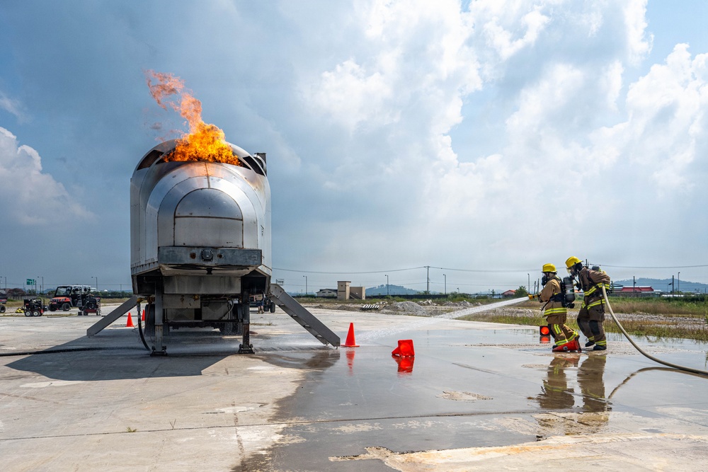 Two firefighters in full protective gear and yellow helmets are conducting a live fire training exercise on a concrete pad. They are directing a hose toward a metal aircraft fuselage simulator with active flames burning from its top. The scene includes safety markers, foam/water on the ground, and support vehicles in the background.