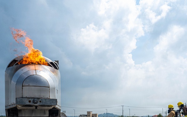Two firefighters in full protective gear and yellow helmets are conducting a live fire training exercise on a concrete pad. They are directing a hose toward a metal aircraft fuselage simulator with active flames burning from its top. The scene includes safety markers, foam/water on the ground, and support vehicles in the background.