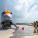 Two firefighters in full protective gear and yellow helmets are conducting a live fire training exercise on a concrete pad. They are directing a hose toward a metal aircraft fuselage simulator with active flames burning from its top. The scene includes safety markers, foam/water on the ground, and support vehicles in the background.