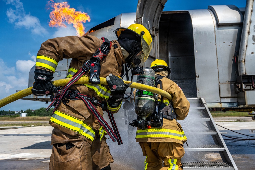 Two firefighters in full protective gear are conducting what appears to be an aircraft firefighting training exercise. They are managing a yellow fire hose near a large aircraft fuselage while an active flame burns in the background. The scene takes place on a tarmac under clear blue skies.