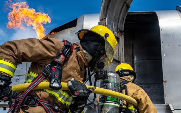 Two firefighters in full protective gear are conducting what appears to be an aircraft firefighting training exercise. They are managing a yellow fire hose near a large aircraft fuselage while an active flame burns in the background. The scene takes place on a tarmac under clear blue skies.