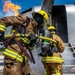 Two firefighters in full protective gear are conducting what appears to be an aircraft firefighting training exercise. They are managing a yellow fire hose near a large aircraft fuselage while an active flame burns in the background. The scene takes place on a tarmac under clear blue skies.