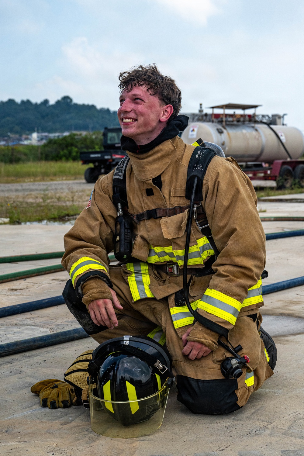 A young firefighter is kneeling on a concrete surface, smiling and appearing to rest after what seems to be a training exercise. He is wearing full turnout gear including a tan/gold structural firefighting jacket with yellow-green reflective striping and an SCBA (Self-Contained Breathing Apparatus) harness on his back. His helmet and gloves are placed on the ground beside him, and his hair and face show signs of heat and exertion.