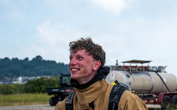 A young firefighter is kneeling on a concrete surface, smiling and appearing to rest after what seems to be a training exercise. He is wearing full turnout gear including a tan/gold structural firefighting jacket with yellow-green reflective striping and an SCBA (Self-Contained Breathing Apparatus) harness on his back. His helmet and gloves are placed on the ground beside him, and his hair and face show signs of heat and exertion.