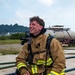 A young firefighter is kneeling on a concrete surface, smiling and appearing to rest after what seems to be a training exercise. He is wearing full turnout gear including a tan/gold structural firefighting jacket with yellow-green reflective striping and an SCBA (Self-Contained Breathing Apparatus) harness on his back. His helmet and gloves are placed on the ground beside him, and his hair and face show signs of heat and exertion.
