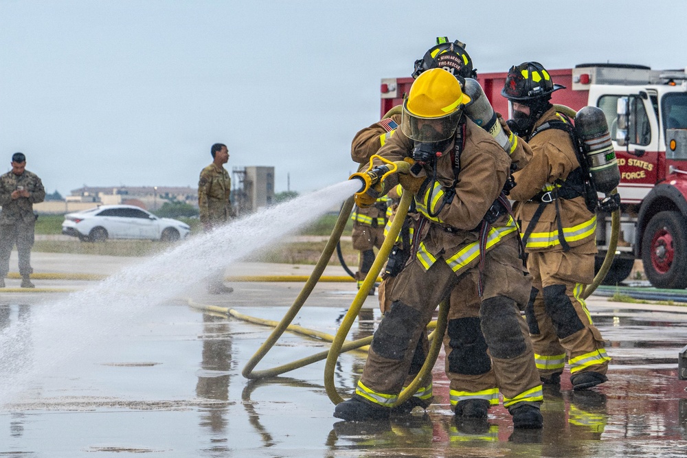 Two firefighters in full protective gear and SCBA (Self-Contained Breathing Apparatus) are operating a fire hose during what appears to be a training exercise at a United States Air Force base. A red fire truck with 'United States Air Force' markings is visible in the background, along with military personnel in camouflage uniforms observing the exercise. The wet pavement and active water stream indicate the drill is in progress.