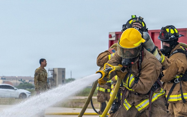 Two firefighters in full protective gear and SCBA (Self-Contained Breathing Apparatus) are operating a fire hose during what appears to be a training exercise at a United States Air Force base. A red fire truck with 'United States Air Force' markings is visible in the background, along with military personnel in camouflage uniforms observing the exercise. The wet pavement and active water stream indicate the drill is in progress.