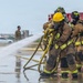 Two firefighters in full protective gear and SCBA (Self-Contained Breathing Apparatus) are operating a fire hose during what appears to be a training exercise at a United States Air Force base. A red fire truck with 'United States Air Force' markings is visible in the background, along with military personnel in camouflage uniforms observing the exercise. The wet pavement and active water stream indicate the drill is in progress.