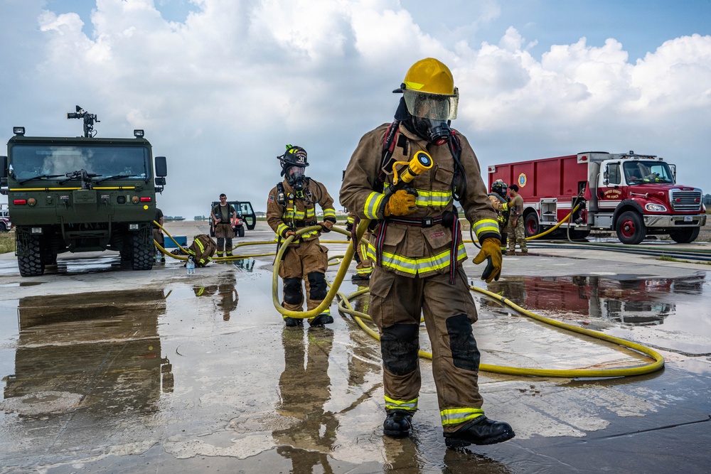 The image shows firefighters in full personal protective equipment (PPE) conducting what appears to be a training exercise on a wet tarmac or airfield. Two primary firefighters in tan/brown turnout gear with yellow reflective stripes are handling yellow fire hoses, with additional personnel and military/fire vehicles visible in the background. The presence of a red fire truck marked 'United States Air Force' and a military green tactical vehicle suggests this is a U.S. Air Force base firefighting drill.