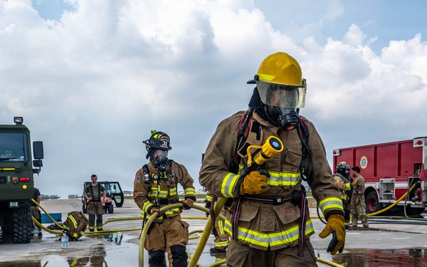 The image shows firefighters in full personal protective equipment (PPE) conducting what appears to be a training exercise on a wet tarmac or airfield. Two primary firefighters in tan/brown turnout gear with yellow reflective stripes are handling yellow fire hoses, with additional personnel and military/fire vehicles visible in the background. The presence of a red fire truck marked 'United States Air Force' and a military green tactical vehicle suggests this is a U.S. Air Force base firefighting drill.