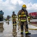 The image shows firefighters in full personal protective equipment (PPE) conducting what appears to be a training exercise on a wet tarmac or airfield. Two primary firefighters in tan/brown turnout gear with yellow reflective stripes are handling yellow fire hoses, with additional personnel and military/fire vehicles visible in the background. The presence of a red fire truck marked 'United States Air Force' and a military green tactical vehicle suggests this is a U.S. Air Force base firefighting drill.