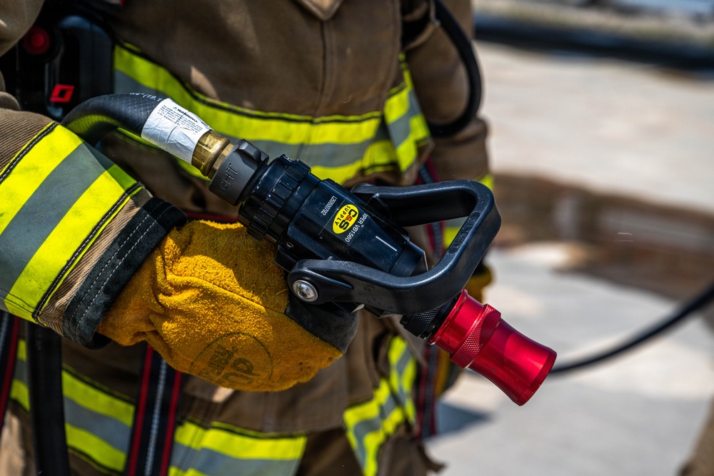 A firefighter in full turnout gear is holding a fire hose nozzle assembly. The image is a close-up shot focusing on the gloved hands gripping a black nozzle with a red tip, connected to a fire hose. The firefighter's gear features the characteristic yellow-green reflective striping on a tan/brown bunker coat.