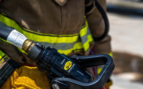 A firefighter in full turnout gear is holding a fire hose nozzle assembly. The image is a close-up shot focusing on the gloved hands gripping a black nozzle with a red tip, connected to a fire hose. The firefighter's gear features the characteristic yellow-green reflective striping on a tan/brown bunker coat.