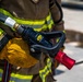 A firefighter in full turnout gear is holding a fire hose nozzle assembly. The image is a close-up shot focusing on the gloved hands gripping a black nozzle with a red tip, connected to a fire hose. The firefighter's gear features the characteristic yellow-green reflective striping on a tan/brown bunker coat.