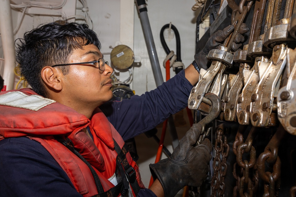 A young man wearing glasses, a navy blue long-sleeve shirt, a red life vest, and black work gloves is handling metal rigging hardware mounted on a wall rack. He appears to be working aboard a vessel, inspecting or retrieving shackles, snap hooks, and chains from a storage rack. The setting appears to be an interior compartment or storage area of a ship.