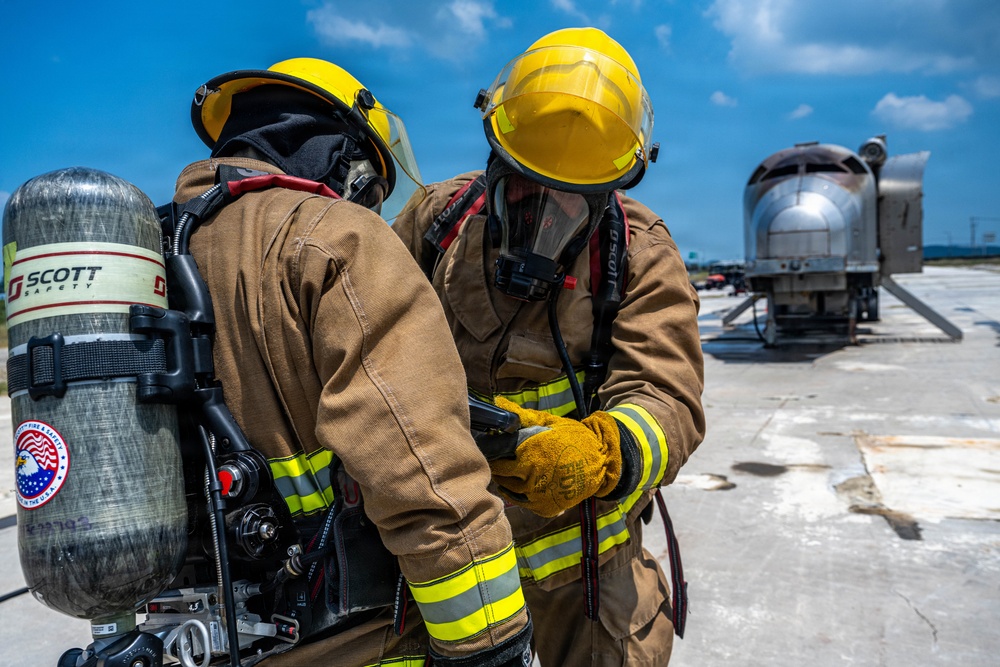 Two firefighters in full personal protective equipment are consulting a handheld device on what appears to be an airfield tarmac. Both are wearing yellow helmets, tan turnout gear with reflective striping, and SCBA (Self-Contained Breathing Apparatus) units branded 'Scott Safety'. A silver aircraft fuselage or training prop is visible in the background, suggesting this may be an aircraft rescue and firefighting (ARFF) training exercise.