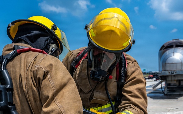 Two firefighters in full personal protective equipment are consulting a handheld device on what appears to be an airfield tarmac. Both are wearing yellow helmets, tan turnout gear with reflective striping, and SCBA (Self-Contained Breathing Apparatus) units branded 'Scott Safety'. A silver aircraft fuselage or training prop is visible in the background, suggesting this may be an aircraft rescue and firefighting (ARFF) training exercise.