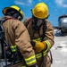 Two firefighters in full personal protective equipment are consulting a handheld device on what appears to be an airfield tarmac. Both are wearing yellow helmets, tan turnout gear with reflective striping, and SCBA (Self-Contained Breathing Apparatus) units branded 'Scott Safety'. A silver aircraft fuselage or training prop is visible in the background, suggesting this may be an aircraft rescue and firefighting (ARFF) training exercise.
