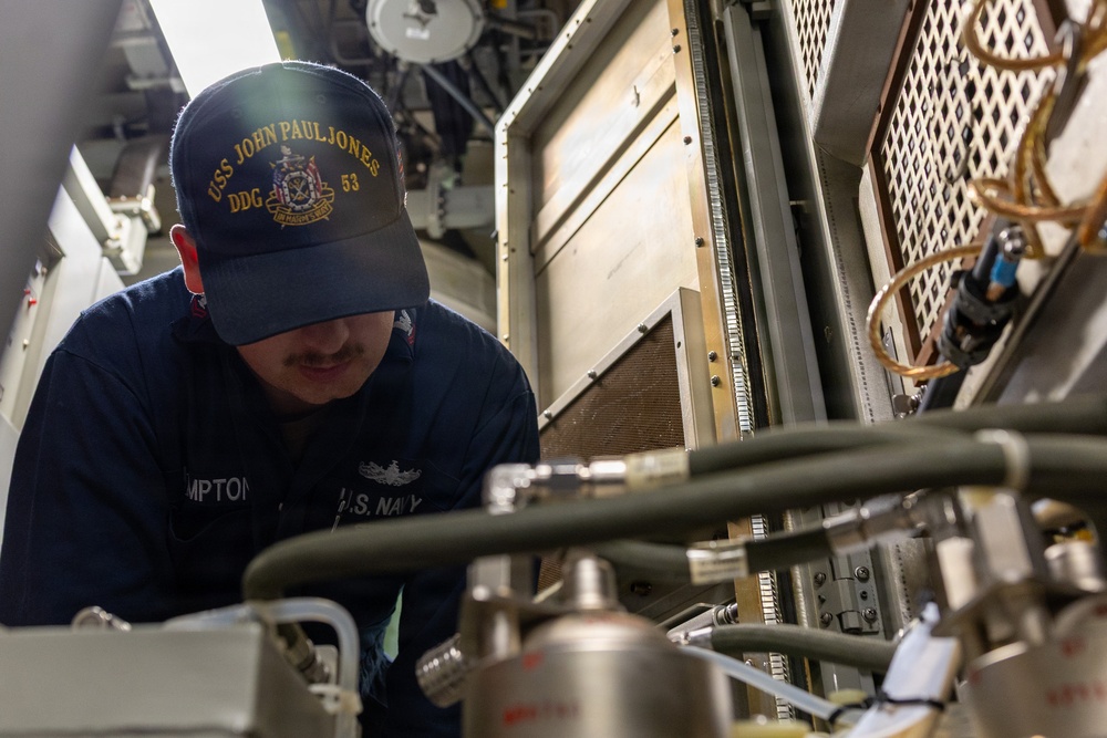 A U.S. Navy sailor assigned to USS John Paul Jones (DDG-53) is performing maintenance or inspection work on shipboard equipment. The sailor is wearing a navy blue coverall uniform and a ship's ball cap, leaning over complex machinery with cables, pipes, and electronic components. The setting appears to be an interior mechanical or electronics space aboard the destroyer.