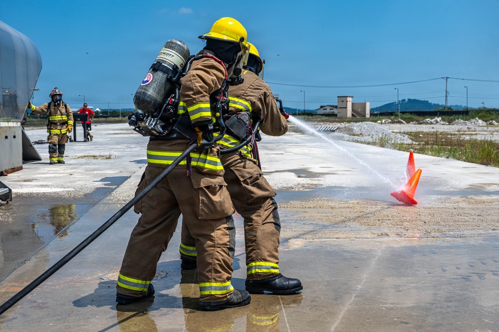 Two firefighters in full turnout gear and SCBA (Self-Contained Breathing Apparatus) are conducting a hose training exercise on an outdoor concrete pad. They are directing a water stream toward an orange traffic cone as a target marker. Additional personnel in firefighting gear are visible in the background near what appears to be a training apparatus.
