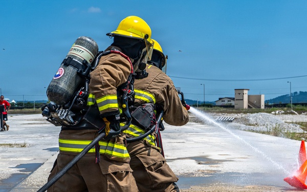 Two firefighters in full turnout gear and SCBA (Self-Contained Breathing Apparatus) are conducting a hose training exercise on an outdoor concrete pad. They are directing a water stream toward an orange traffic cone as a target marker. Additional personnel in firefighting gear are visible in the background near what appears to be a training apparatus.