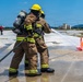 Two firefighters in full turnout gear and SCBA (Self-Contained Breathing Apparatus) are conducting a hose training exercise on an outdoor concrete pad. They are directing a water stream toward an orange traffic cone as a target marker. Additional personnel in firefighting gear are visible in the background near what appears to be a training apparatus.