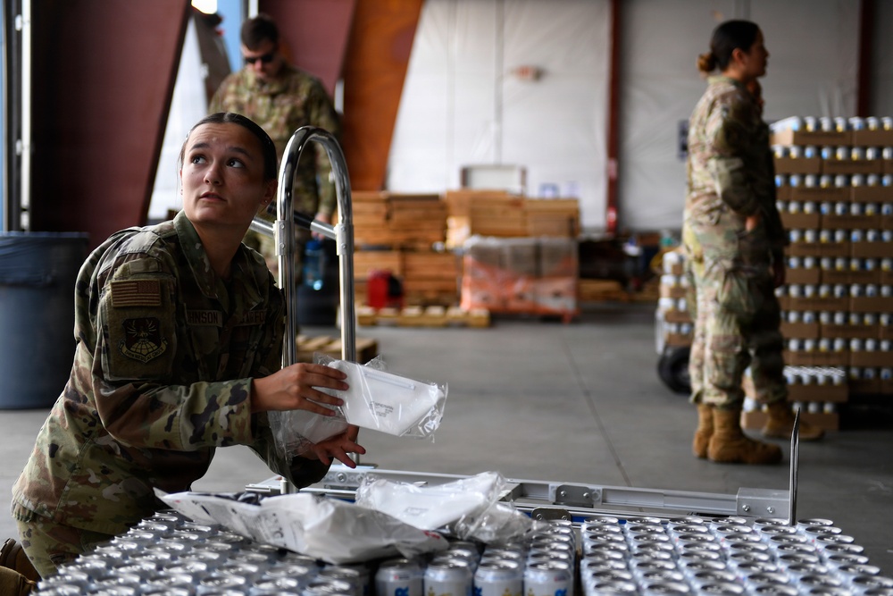 Three military personnel in US Army/Air Force OCP (Operational Camouflage Pattern) uniforms are working in what appears to be a warehouse or hangar facility. The foreground soldier is handling packaged supplies on a metal cart, while two others manage stacked inventory in the background. The scene suggests a logistics, supply, or humanitarian aid operation.