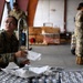 Three military personnel in US Army/Air Force OCP (Operational Camouflage Pattern) uniforms are working in what appears to be a warehouse or hangar facility. The foreground soldier is handling packaged supplies on a metal cart, while two others manage stacked inventory in the background. The scene suggests a logistics, supply, or humanitarian aid operation.