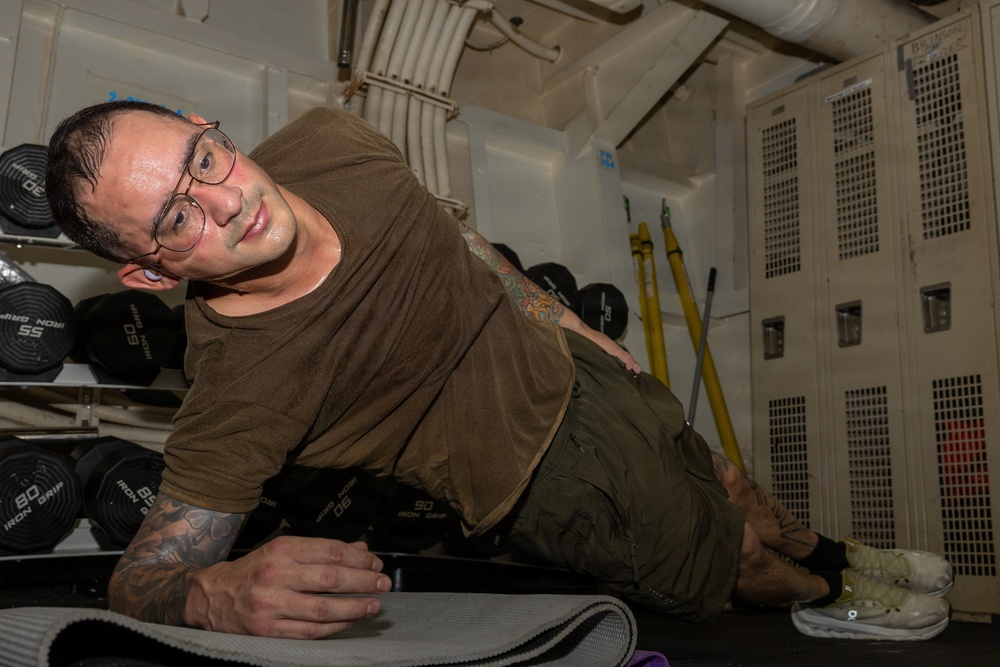 A man wearing glasses and a brown/olive t-shirt is performing a side plank exercise on a yoga mat in what appears to be a shipboard gym or fitness area. He has visible tattoos on both arms and is wearing olive/green cargo pants and light-colored athletic shoes. The setting includes a dumbbell rack with Iron Grip weights, metal lockers, and yellow cleaning equipment in the background.